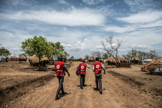 Three men walking in an open space surrounding by dirt paths and huts 