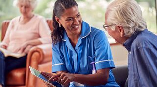 Healthcare practitioner holding a tablet is smiling whilst sitting with elderly man