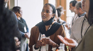 Close up of person in brown jacket and black scarf, talking with a group of colleagues