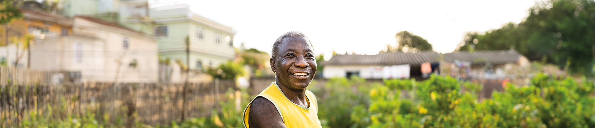 Smiling older person standing amongst crops, holding a wooden gardening tool