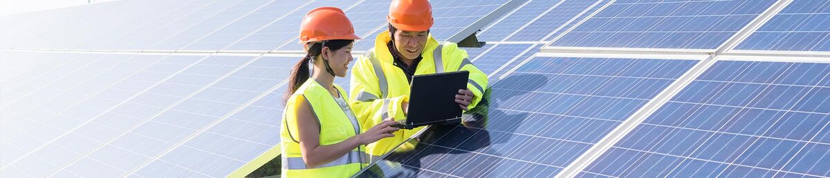 Two people in yellow hi-vis vests and orange safety helmets leaning on a solar panel in a field, looking at an iPad