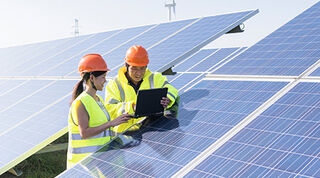 Two people in yellow hi-vis vests and orange safety helmets leaning on a solar panel in a field, looking at an iPad