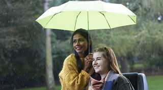 Woman holding a green umbrella over another woman in a wheelchair, both smiling out to the left